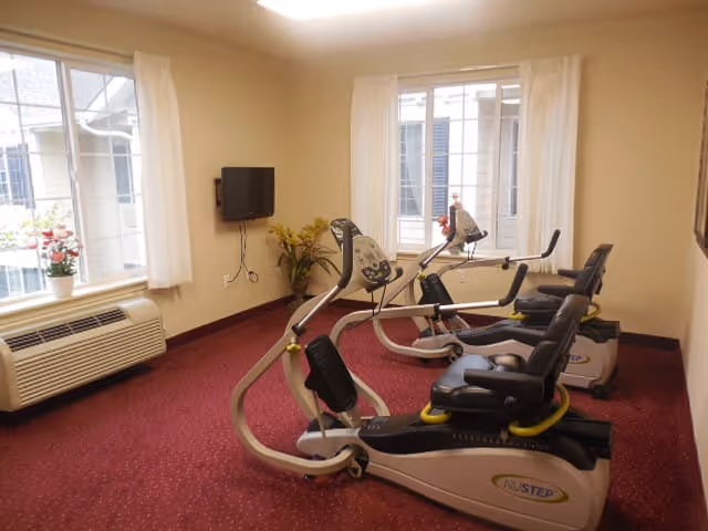 A small exercise room with two recumbent stationary bikes facing a wall-mounted TV. The room has beige walls, red carpet, two windows with white curtains, and a white air conditioning unit below one window. There are also some potted plants near the TV and windows.