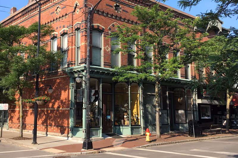 Two-story red brick corner commercial building with green-trimmed storefront windows, trees along the sidewalk, and a yellow fire hydrant.
