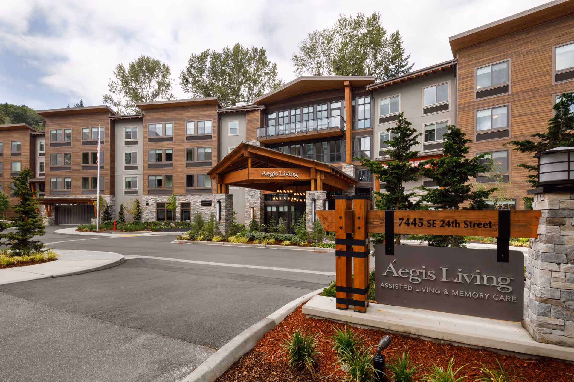 Exterior view of Aegis Living Mercer Island, a multi-story assisted living and memory care facility with a modern design featuring wood and stone accents. The entrance has a covered driveway and a sign displaying the address 7445 SE 24th Street and the facility name.