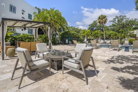 Outdoor patio area with cushioned chairs arranged around a fire pit table, a shaded seating area with a canopy, and several lounge chairs near a pool, surrounded by palm trees and greenery under a blue sky with some clouds.