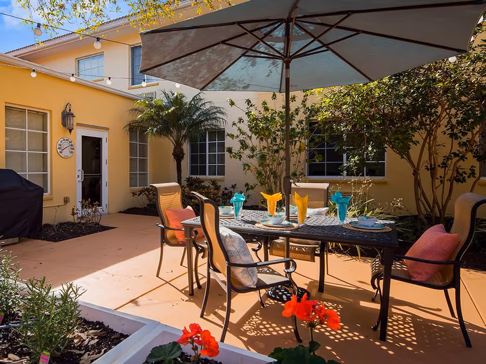 Outdoor patio area with a table and four chairs under a large umbrella. The table is set with colorful napkins and glassware. Surrounding the patio are plants, bushes, and a yellow building with windows and a door. String lights hang above the patio.