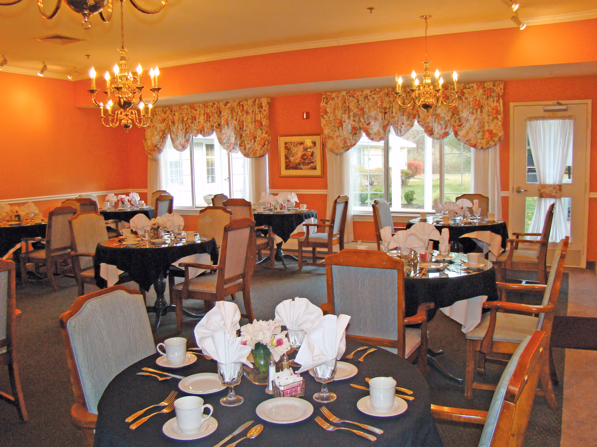 A dining room with multiple round tables covered with black tablecloths and set with white plates, cups, silverware, and folded white napkins. The room has orange walls, floral curtains on the windows, and two chandeliers hanging from the ceiling. Chairs with wooden frames and upholstered seats surround the tables. A door with a window and curtain is visible on the right side.