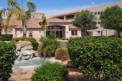 Exterior view of a retirement home building with a tiled roof, surrounded by palm trees, bushes, and a small water feature with rocks in the foreground under a clear blue sky.