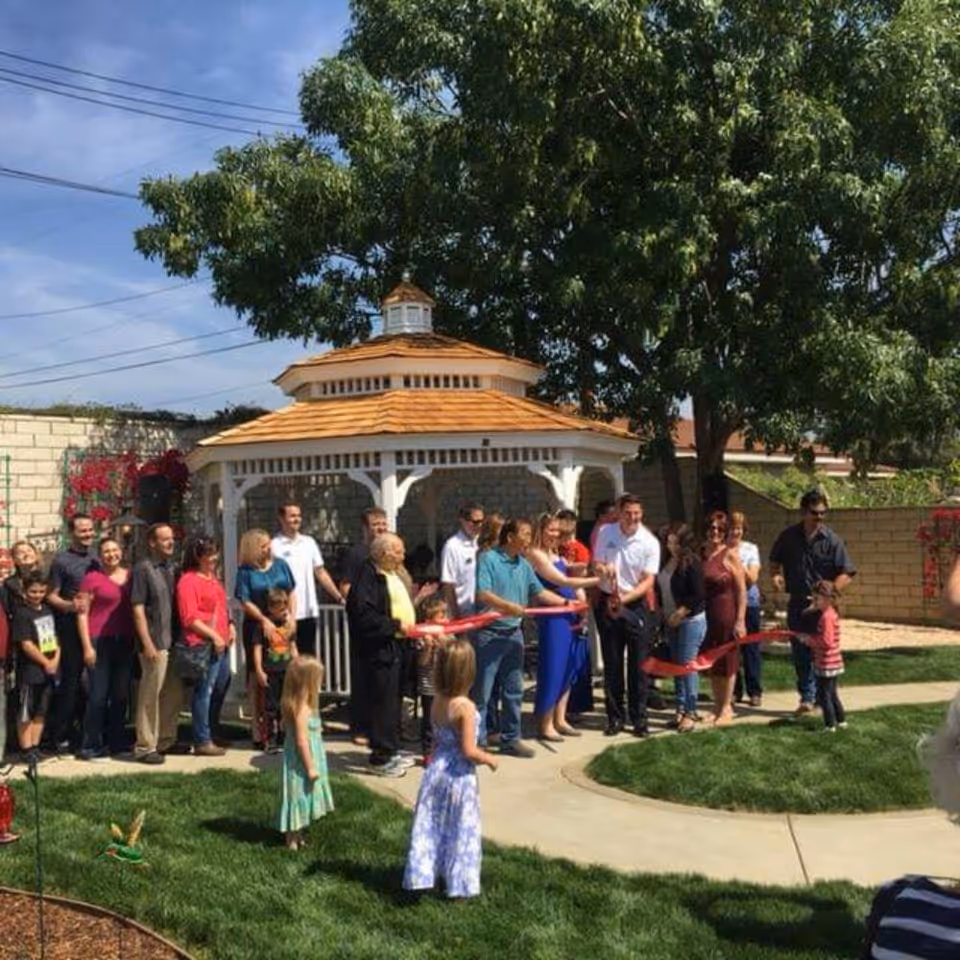 A group of people gathered outdoors in a garden area with a white gazebo and large tree. Several adults and children are present, with some adults holding a red ribbon for a ribbon-cutting ceremony. The scene is sunny with a clear blue sky.
