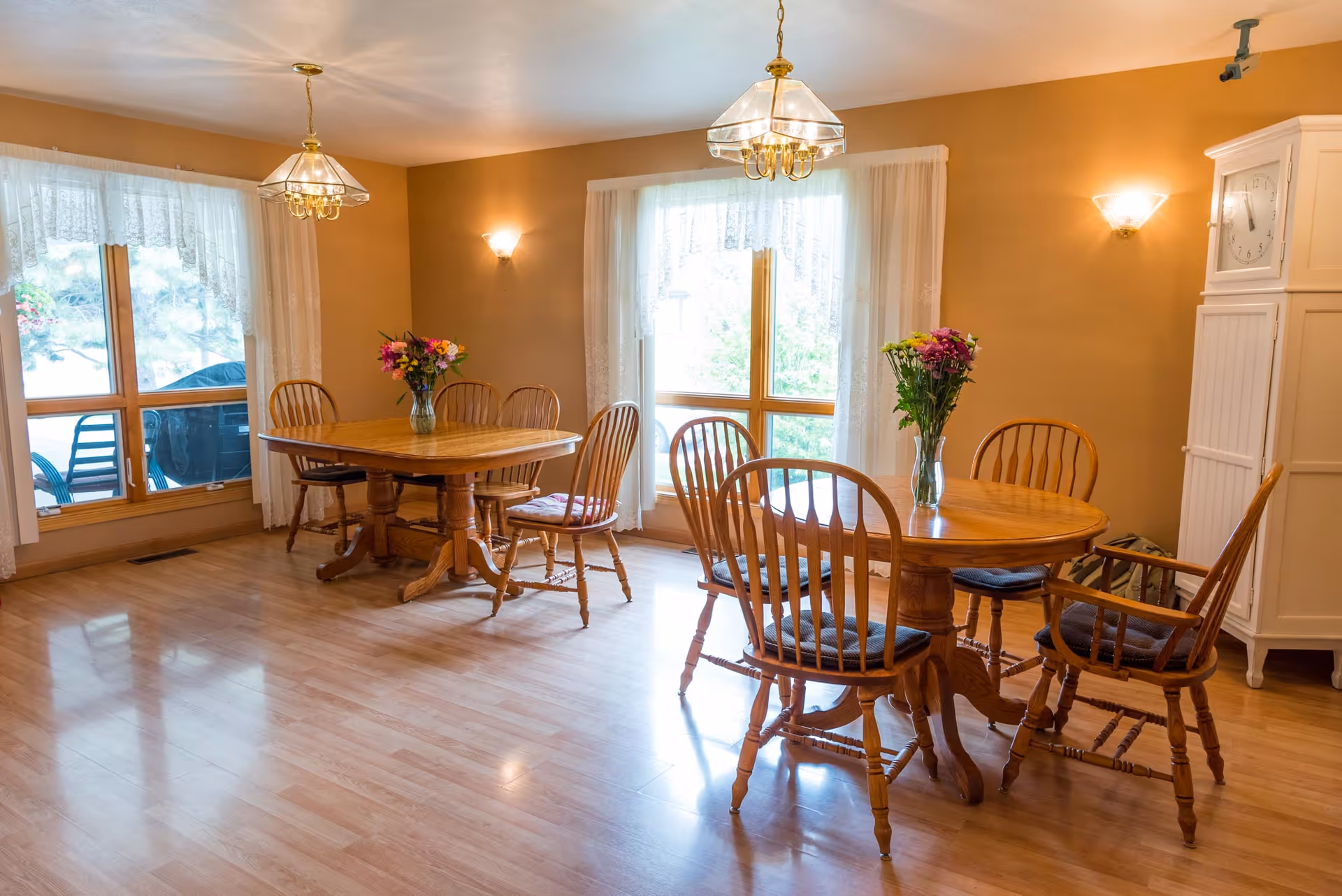 Bright dining room with two wooden tables and matching chairs, vases of flowers, large windows with sheer curtains, and hardwood floors.