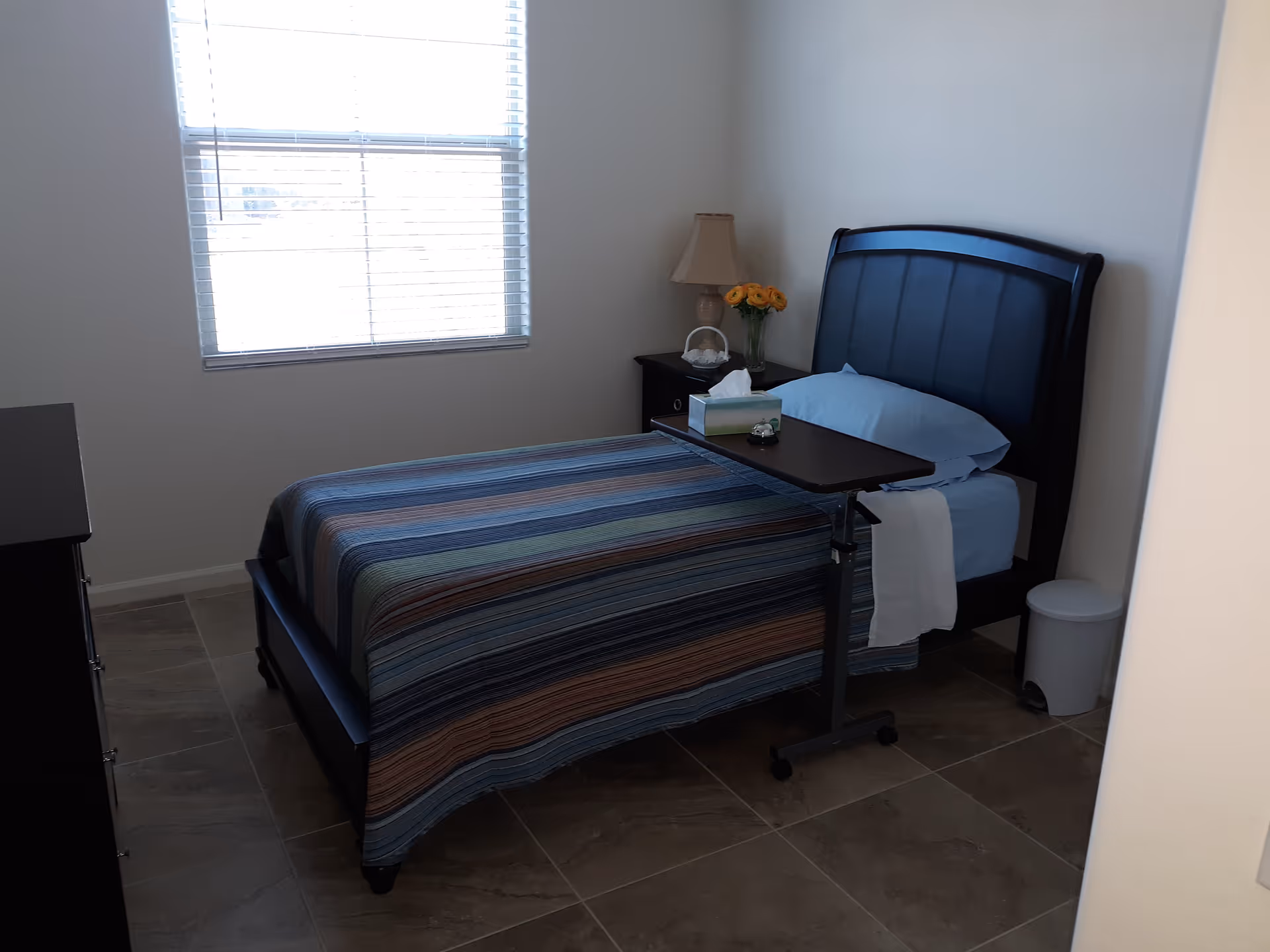 Small, tidy bedroom with a single bed featuring a striped bedspread, an overbed table, bedside lamp and a window.