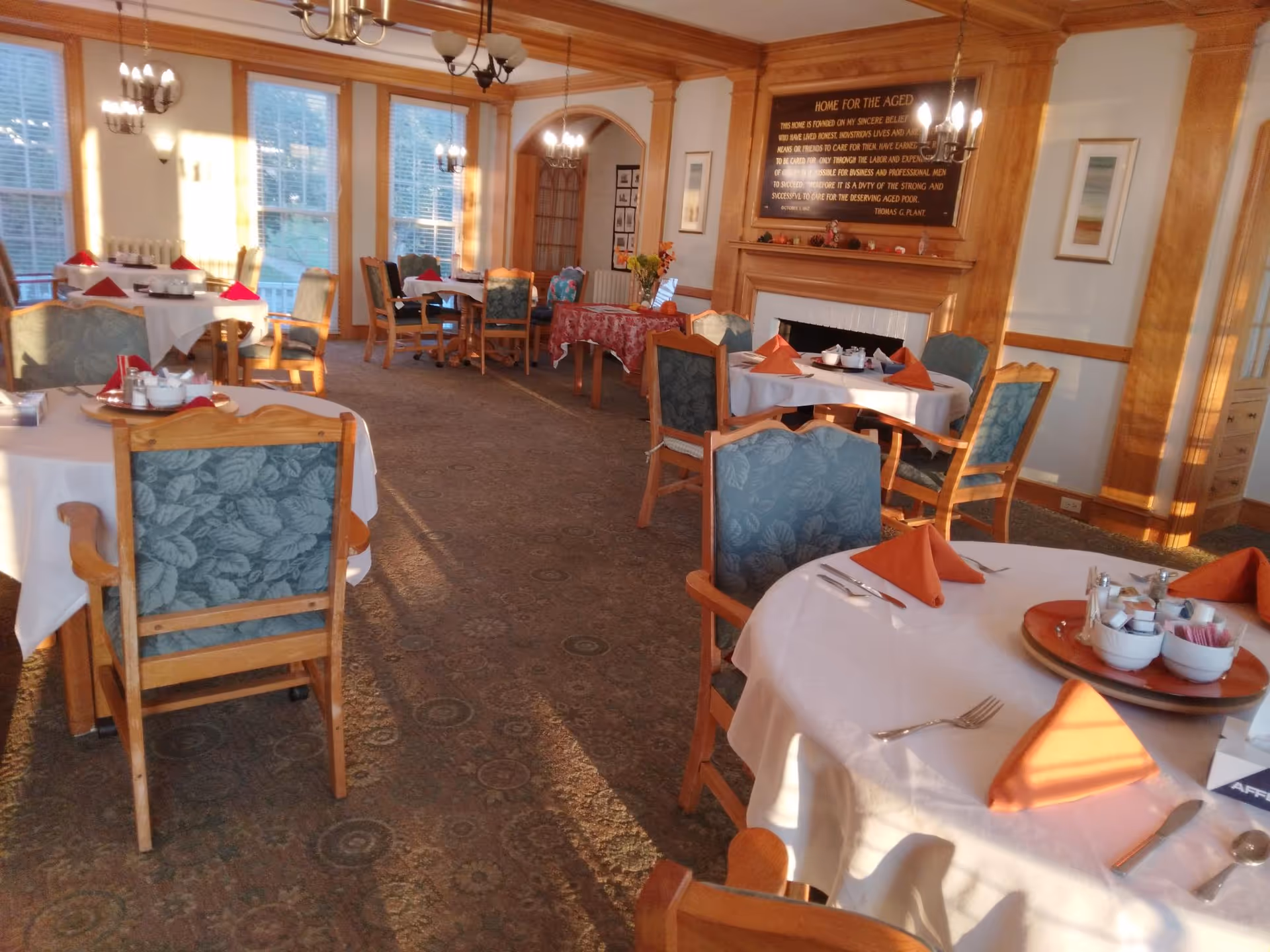 A dining room with several round tables covered with white tablecloths and set with orange folded napkins, silverware, and condiment trays. The room has wooden paneling, large windows letting in natural light, chandeliers, and a carpeted floor. A wooden fireplace with a plaque above it is visible in the background.