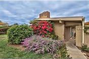 Front entrance of a single-story senior living building with a walkway and flowering shrubs under a cloudy sky.
