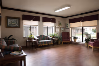 A spacious living room with wooden flooring, several upholstered chairs and sofas arranged around the room, potted plants on small tables, large windows with blinds and valances allowing natural light to enter, and framed artwork on the walls.