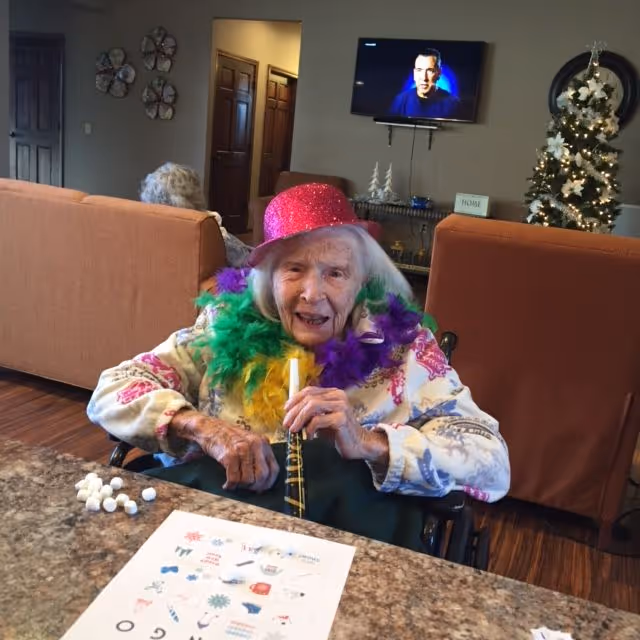 An elderly woman in a pink hat and colorful feather boa sits at a table holding a party horn in a decorated communal living room.
