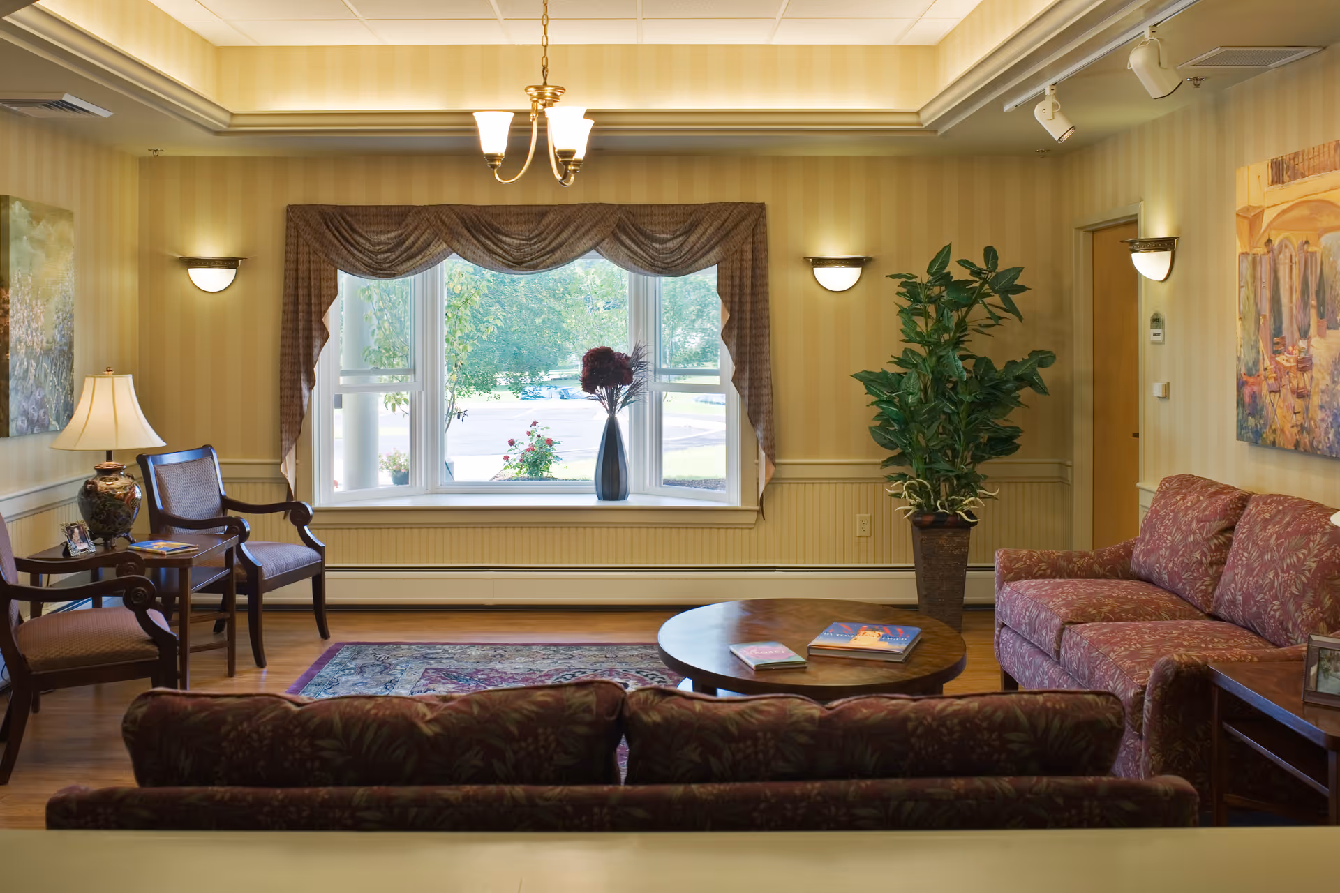 Sunlit common living room with upholstered sofas and chairs, a round coffee table, potted plant, and a large window overlooking greenery.
