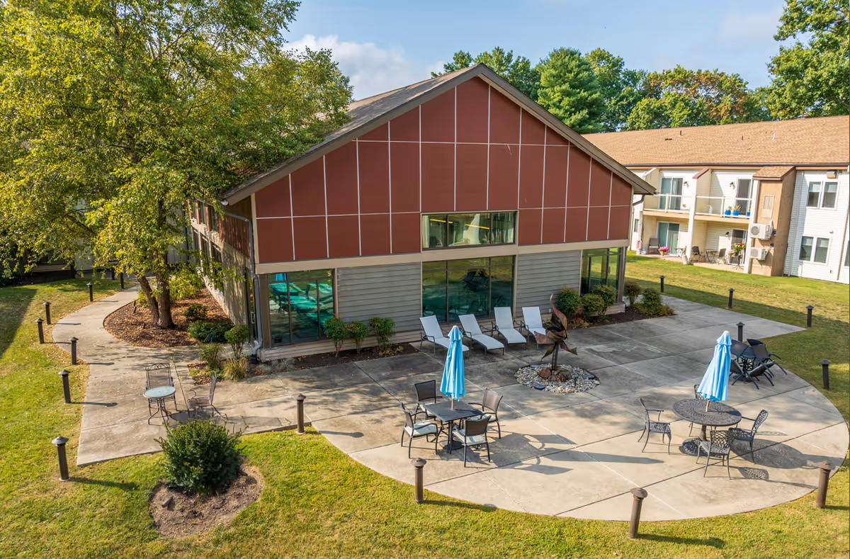 Outdoor patio with round tables, blue umbrellas, lounge chairs and a two-story building surrounded by grass and trees.