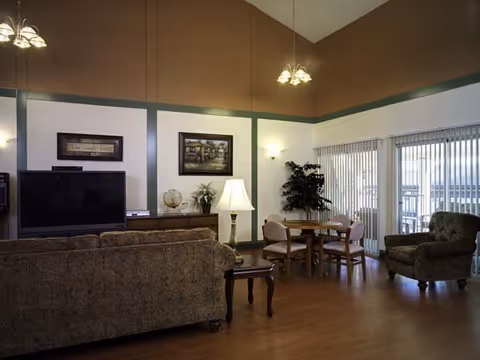 A cozy living room area with a patterned sofa facing a flat-screen TV on a wooden cabinet. There is a table lamp on a side table next to the sofa, a round wooden dining table with four chairs, and a comfortable armchair near large windows with vertical blinds. The walls are painted in two tones with a green trim, and the ceiling has hanging light fixtures.