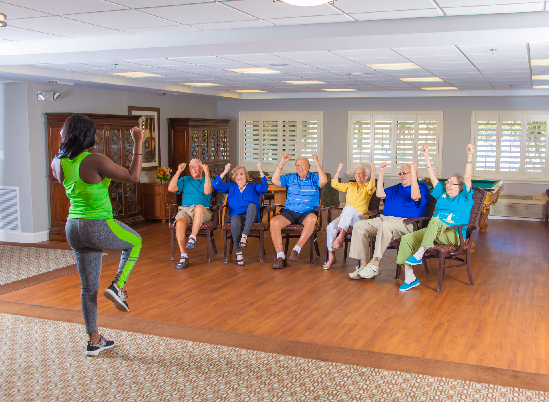 A fitness instructor in a bright green and gray outfit leads a seated exercise class for six elderly individuals in a spacious, well-lit room with wooden flooring and large windows with white shutters. The elderly participants are seated in chairs, raising their arms enthusiastically, following the instructor's movements.