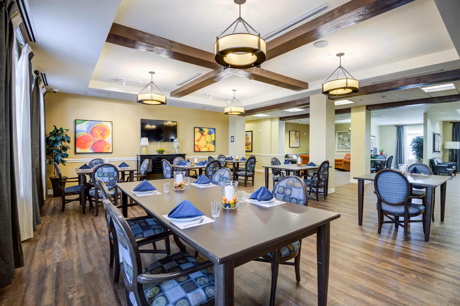 A spacious dining room with multiple tables and chairs arranged neatly. Each table is set with blue napkins, glasses, and small decorative centerpieces. The room features wooden flooring, beige walls adorned with colorful fruit paintings, and large windows with curtains allowing natural light. Ceiling lights with wooden beams add a warm ambiance. In the background, there is a television mounted on the wall and a sideboard with decorative items.