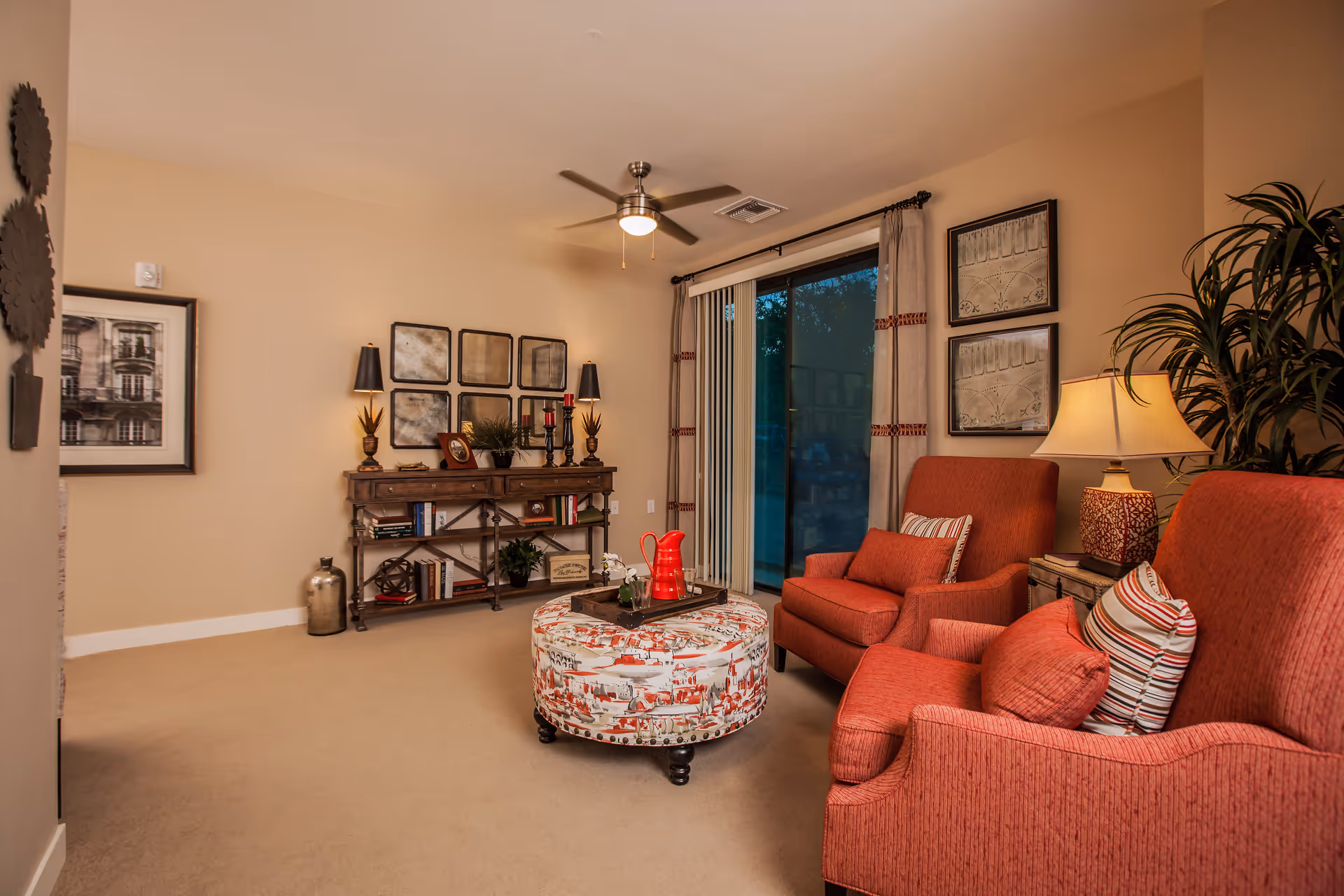 Cozy living room with two red armchairs, a patterned ottoman, and shelving against a beige wall.
