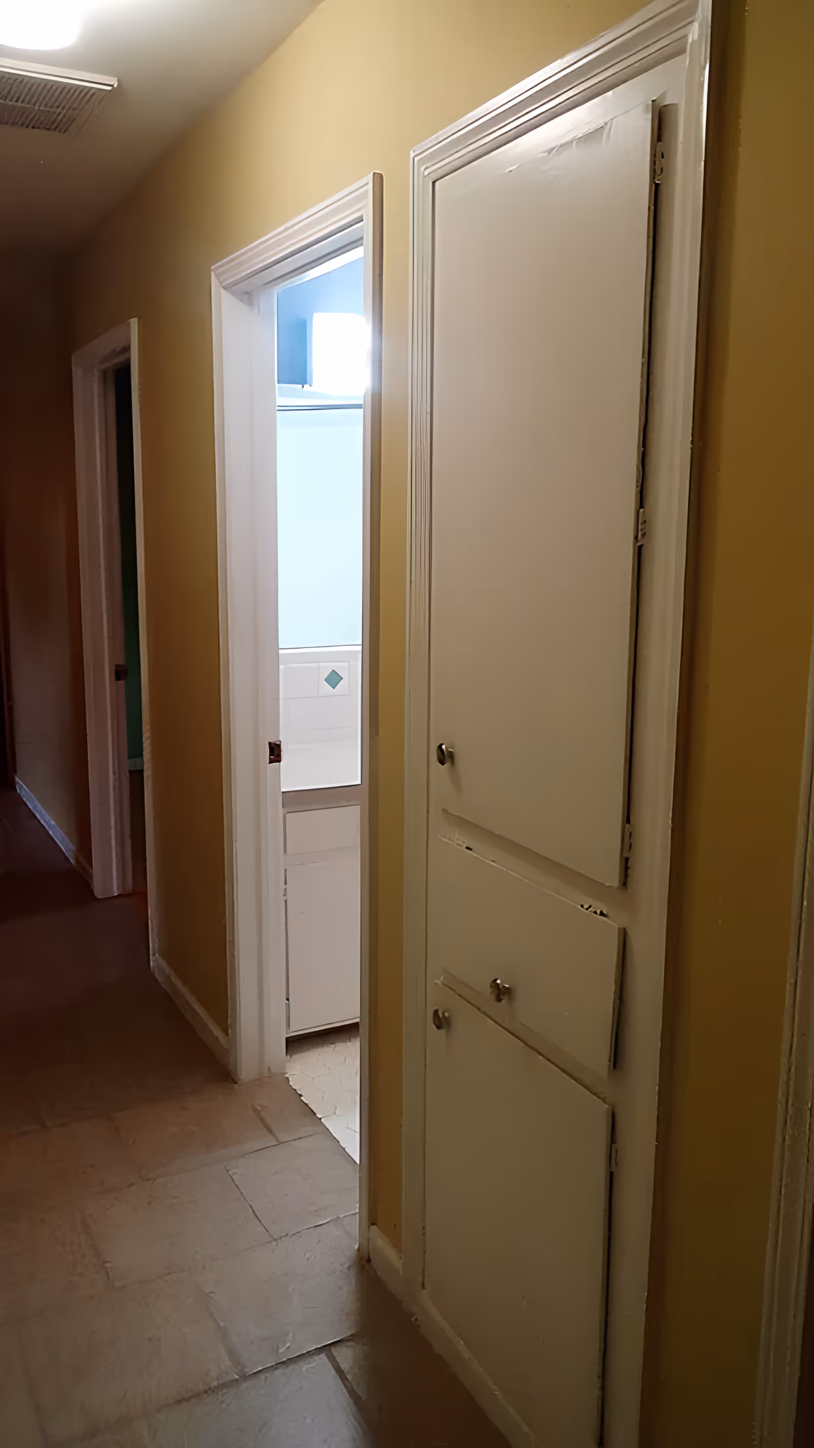 Narrow interior hallway with yellow walls, built-in white linen cabinets on the right and an open doorway revealing a bathroom sink.