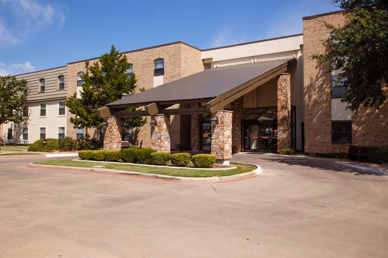 Exterior view of a senior living facility building with a covered entrance supported by stone pillars, surrounded by trimmed bushes and trees under a clear blue sky.