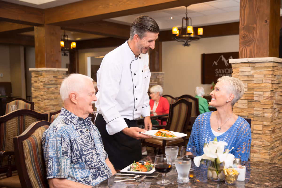 A chef serving a plated meal to an elderly woman seated at a dining table in a restaurant setting. An elderly man is also seated at the table, smiling. The table has glasses of water, wine, and a flower centerpiece. The background shows other diners and warm lighting with wooden beams and stone pillars.