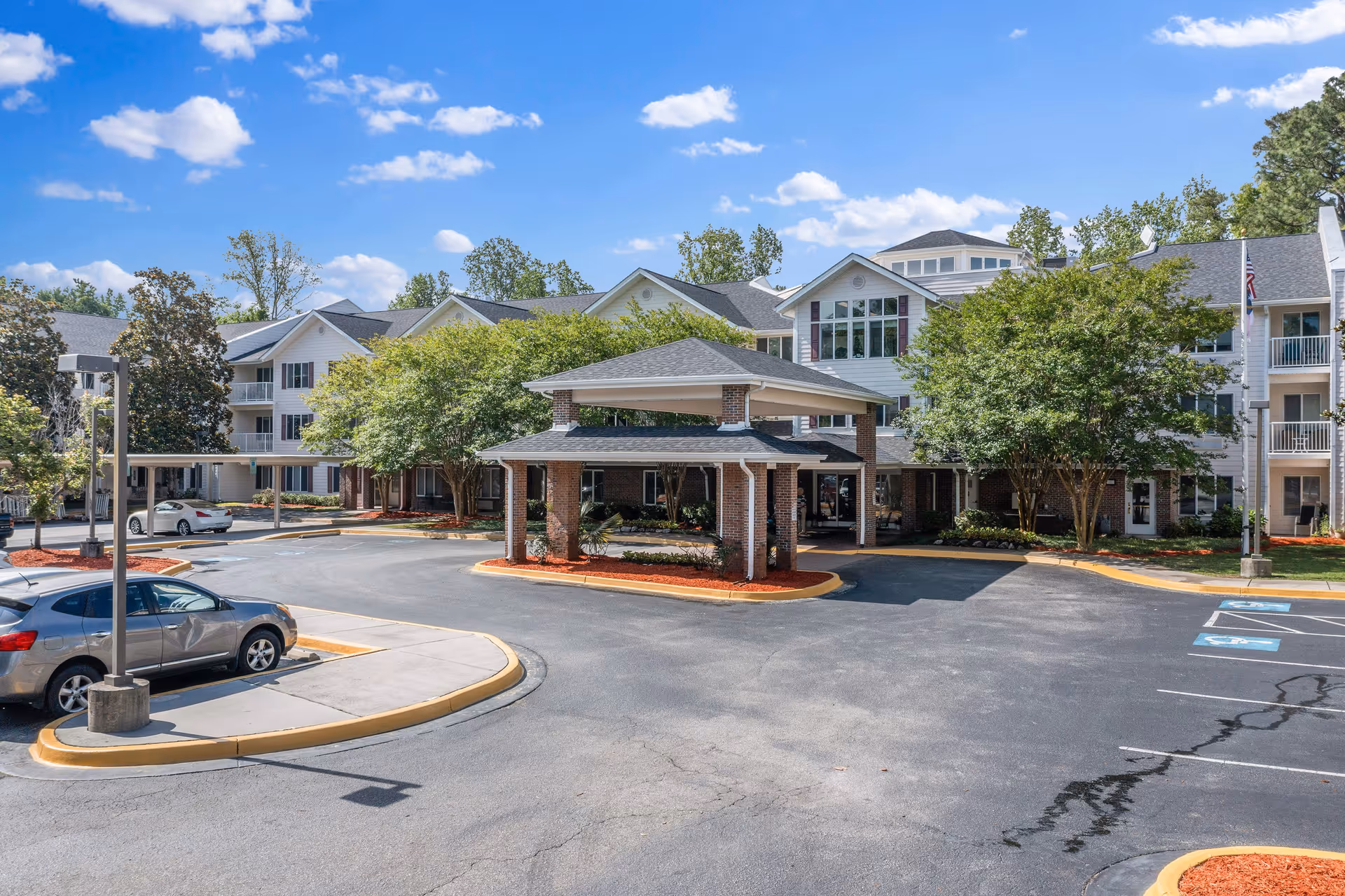 Exterior view of The Woods at Holly Tree Gracious Retirement Living facility showing a multi-story building with white siding and large windows, surrounded by trees and landscaping. There is a covered entrance with brick pillars and a driveway with parked cars and handicap parking spaces under a clear blue sky.