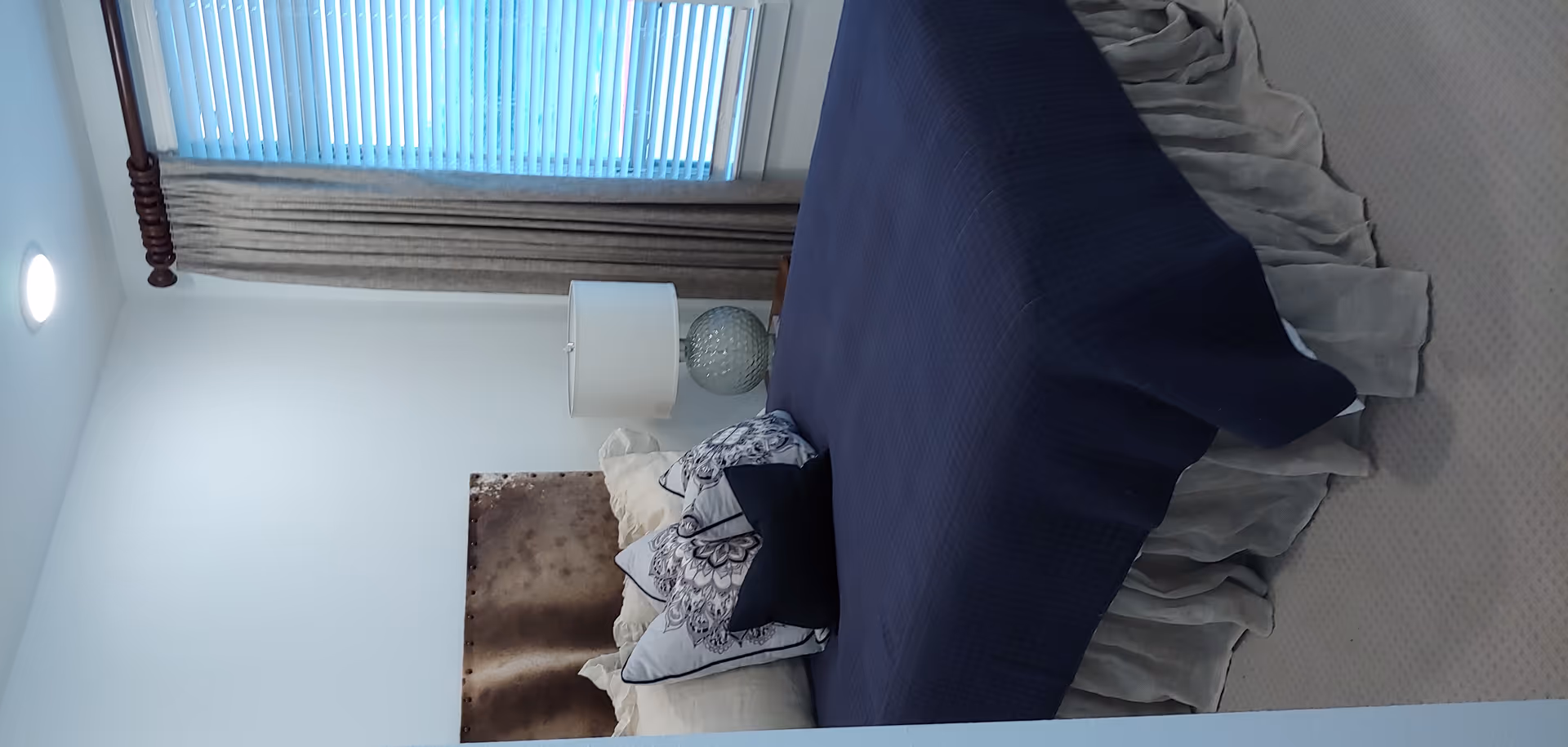 A bedroom with a bed dressed in a navy coverlet and decorative pillows, a bedside lamp, and a window with vertical blinds and curtains.
