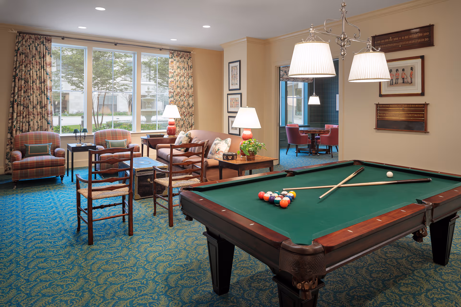 A cozy senior living common room with a pool table in the foreground and a seating area with chairs and lamps by large windows.