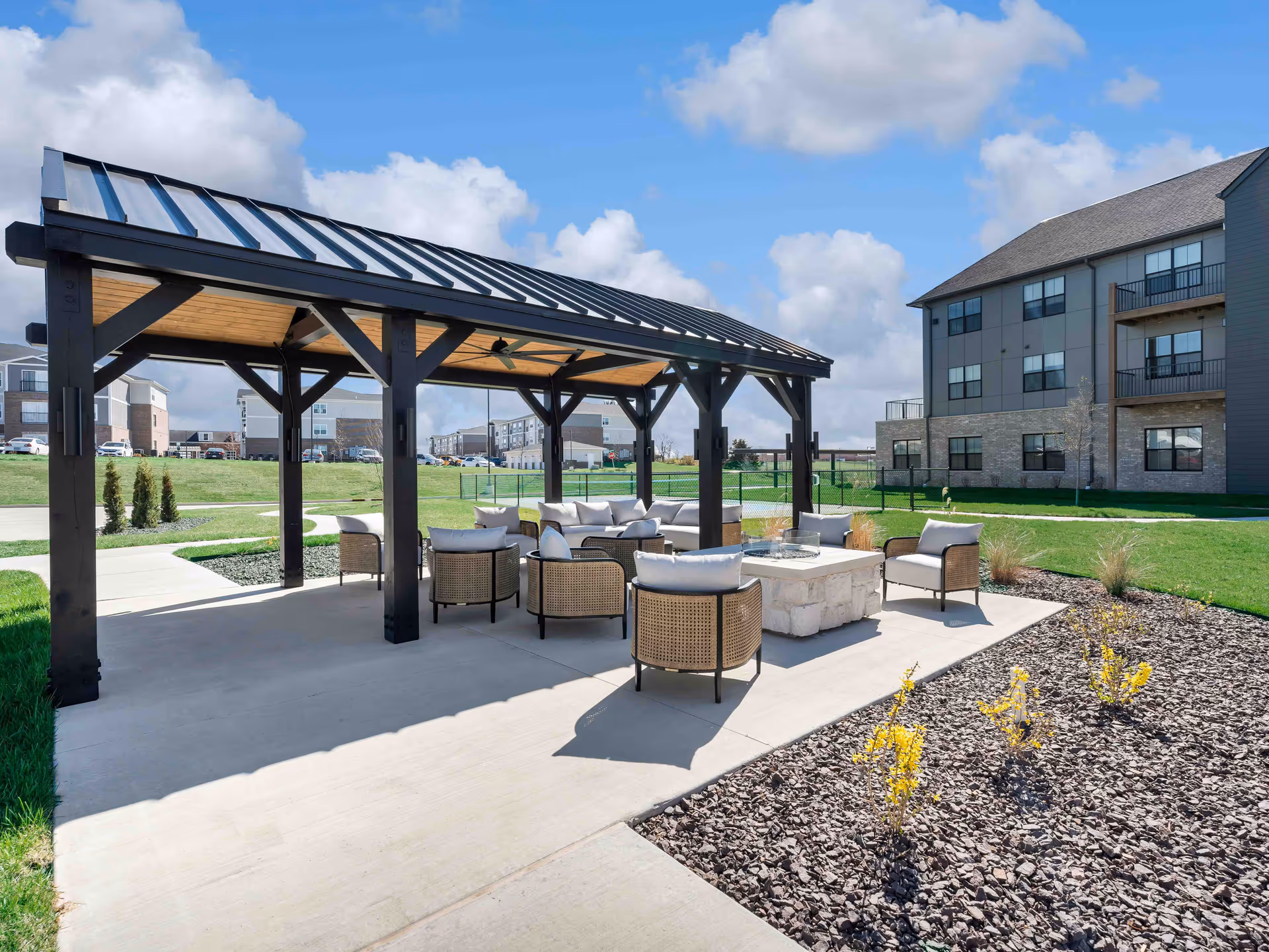 Outdoor seating area at Cedarhurst Senior Living of Wentzville featuring a covered pavilion with a metal roof, cushioned chairs arranged around a stone fire pit, and landscaped surroundings with grass and small plants under a partly cloudy sky.