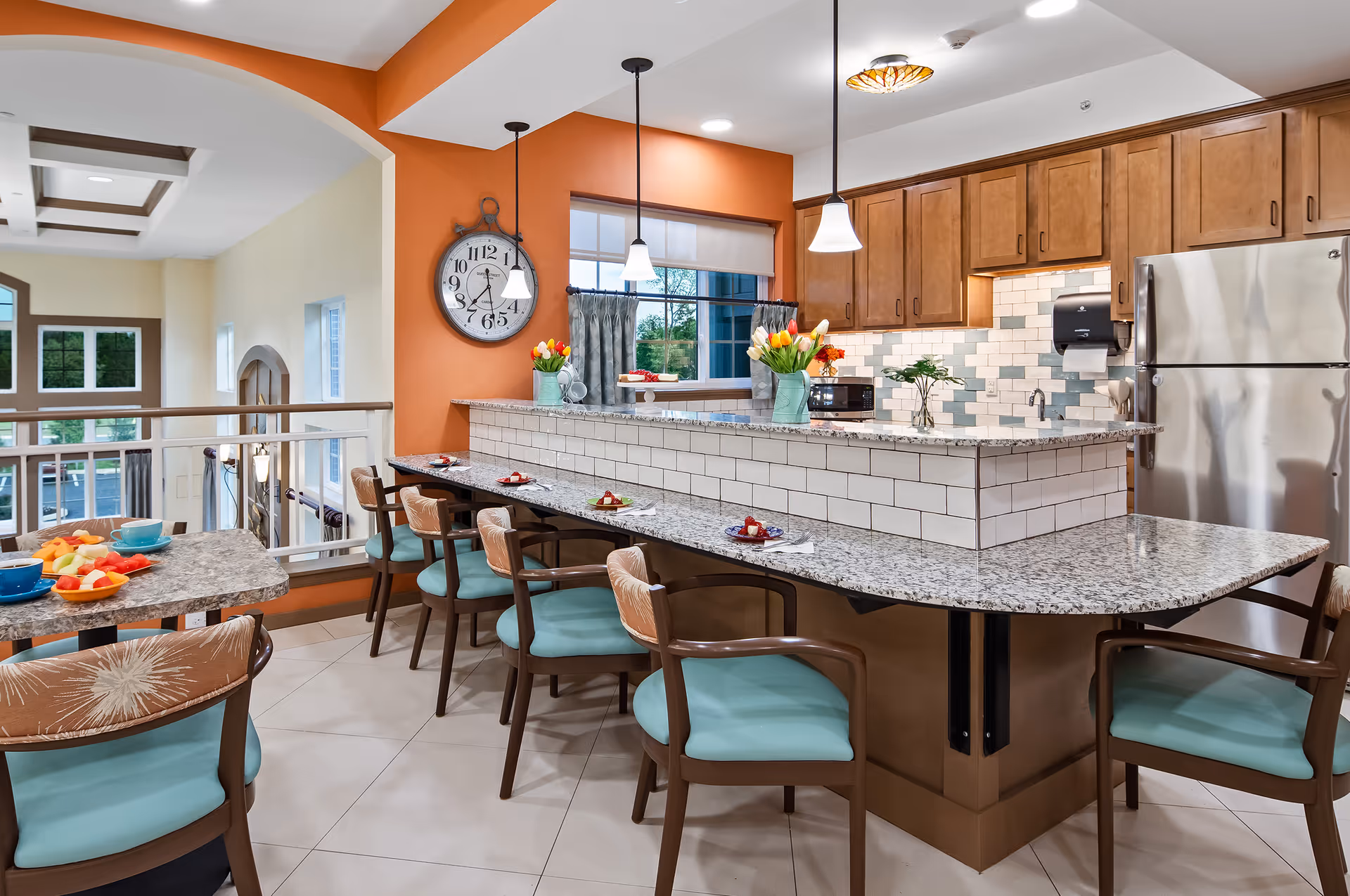 A bright and modern kitchen and dining area with a granite countertop bar featuring six chairs with teal cushions. The kitchen has wooden cabinets, a stainless steel refrigerator, and a tiled backsplash in white and light blue. There are pendant lights hanging above the bar, a large wall clock, and vases with colorful tulips on the counter. A table with bowls of fruit is visible in the foreground, and large windows provide natural light.