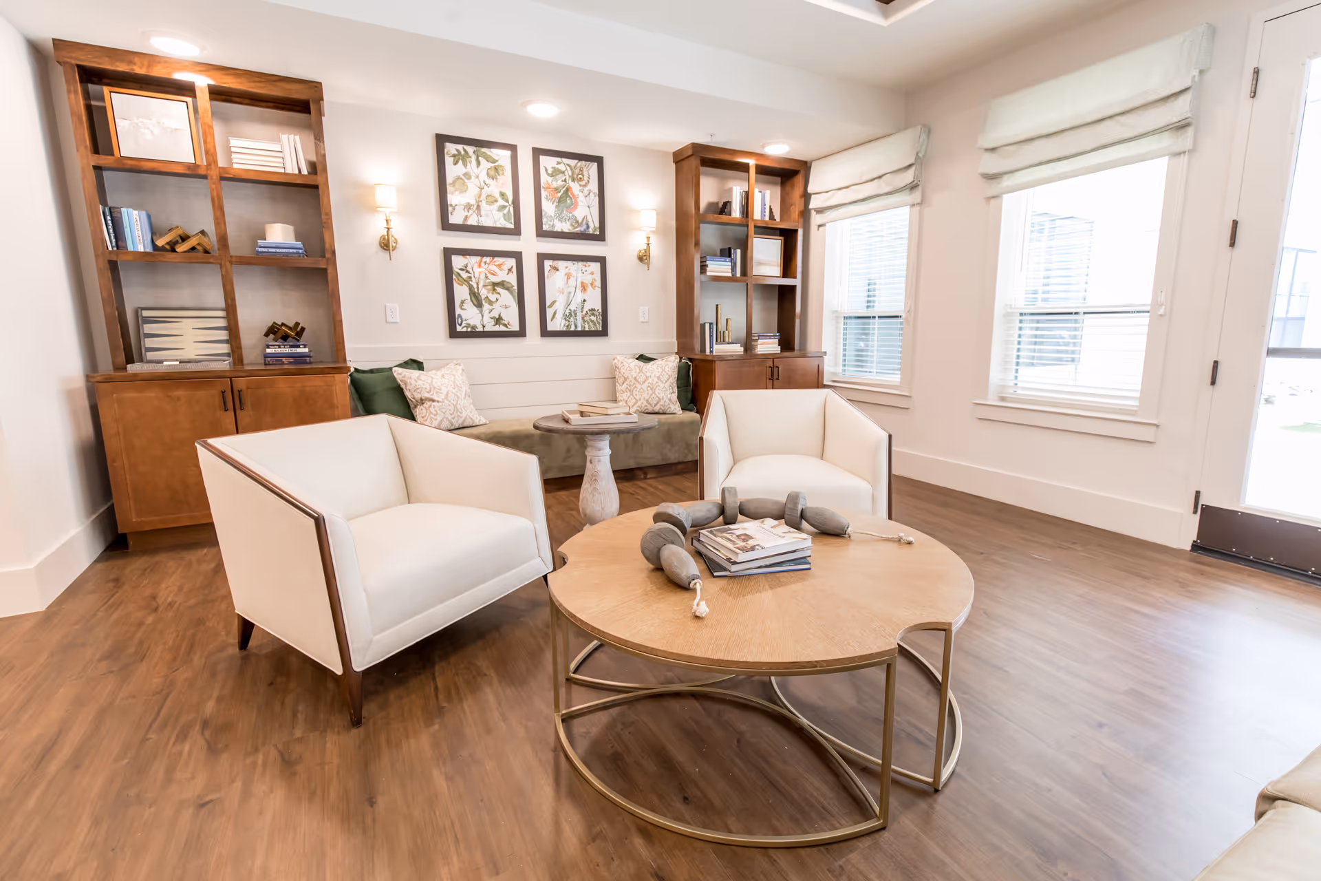 A bright and cozy living room area with two white armchairs, a round wooden coffee table with decorative items and books, a green cushioned bench with pillows against the wall, two wooden bookshelves with books and decor, and four framed botanical prints on the wall. The room has wooden flooring, white walls, and large windows with white blinds.