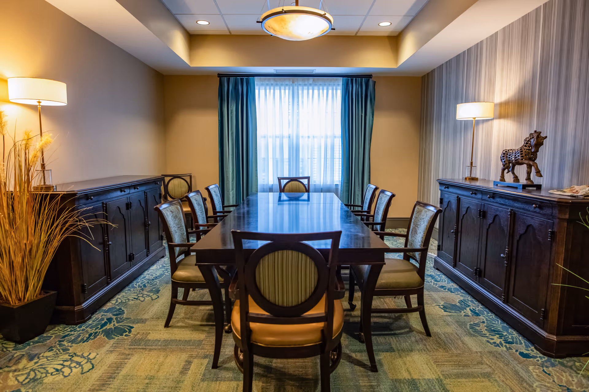 A formal dining/meeting room with a long wooden table surrounded by chairs, sideboards, lamps, and a curtained window.