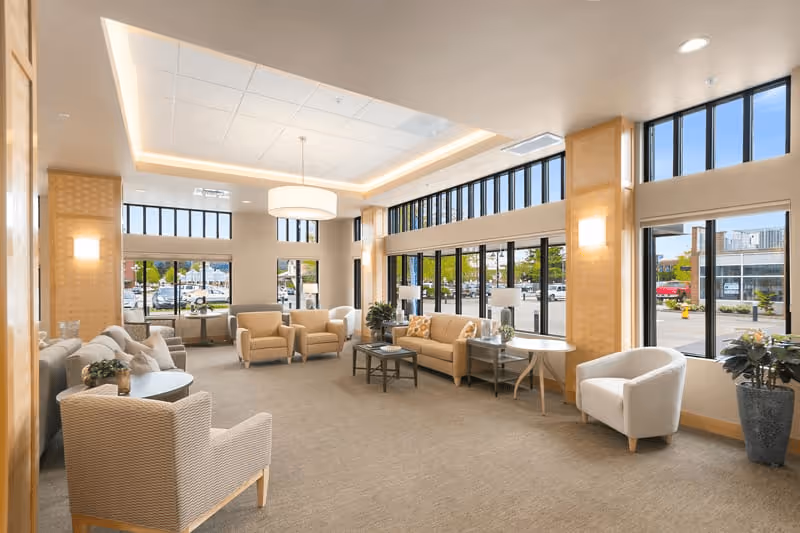 A spacious and well-lit living room area with large windows allowing natural light to fill the space. The room features multiple beige and cream-colored armchairs and sofas arranged around small tables. There are plants placed near the windows and on tables, with modern light fixtures on the ceiling and walls. Outside the windows, a parking lot and some buildings are visible.