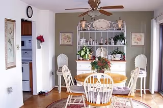 Dining area with a round wooden table and four chairs, a decorative hutch with plants and framed art, and a ceiling fan.