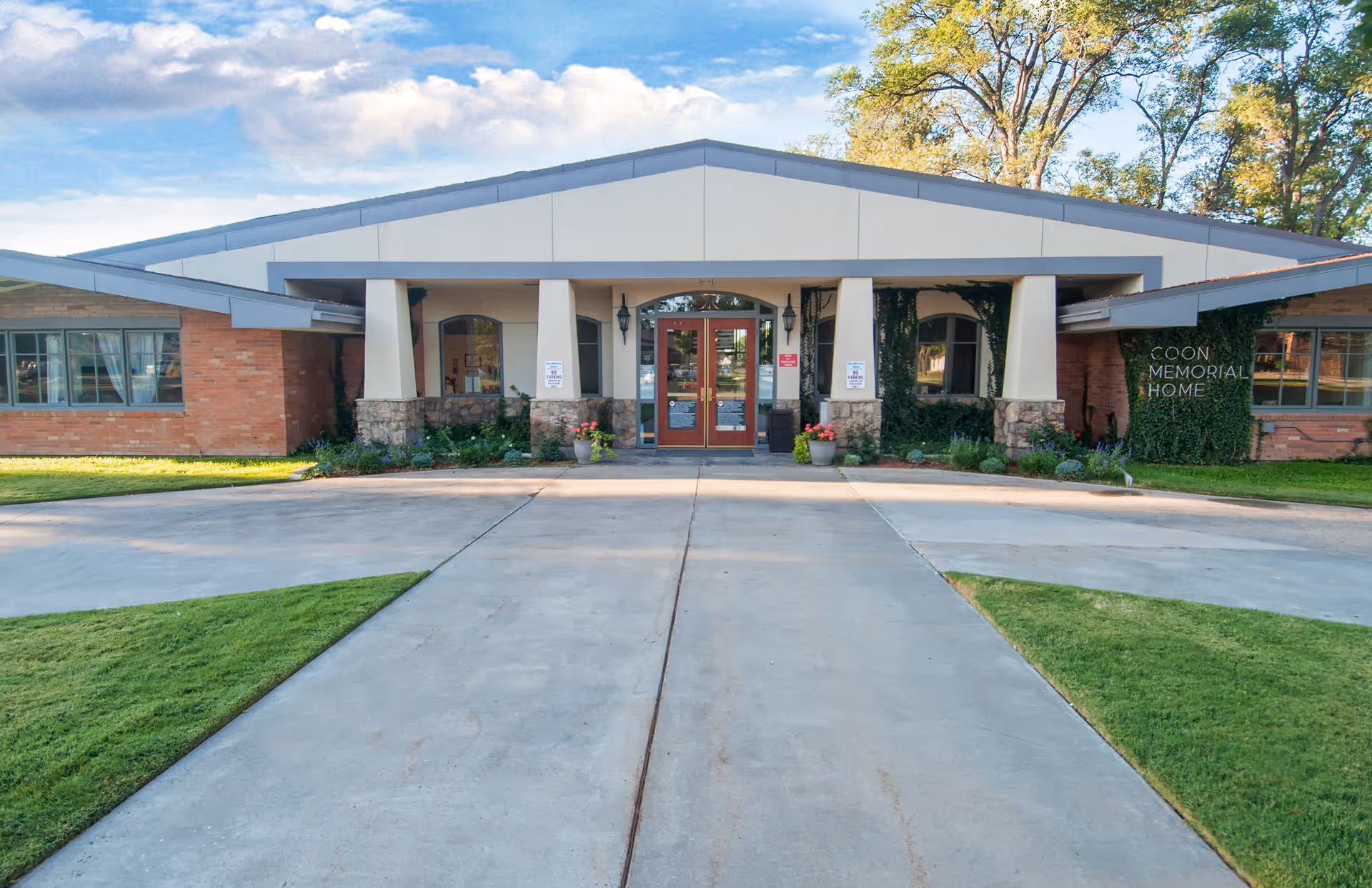 Front exterior view of Coon Memorial Nursing Home building with a wide concrete driveway leading to double glass doors entrance. The building has a combination of brick and light-colored walls with large windows and some greenery around the entrance.
