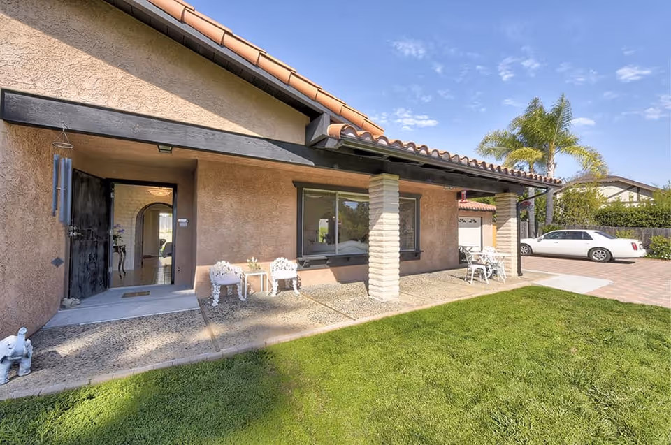Front exterior of a single-story home with a covered porch, patio seating, lawn and a driveway with a white car.