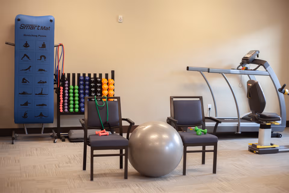 Exercise room with two chairs, a large stability ball, hand weights, resistance bands, a mat rack, and a treadmill against a beige wall.