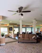 A spacious and well-lit living room area in an assisted living facility featuring a ceiling fan, a brown upholstered sofa with pillows, multiple dining tables and chairs in the background, large windows letting in natural light, and soft beige walls with white trim.