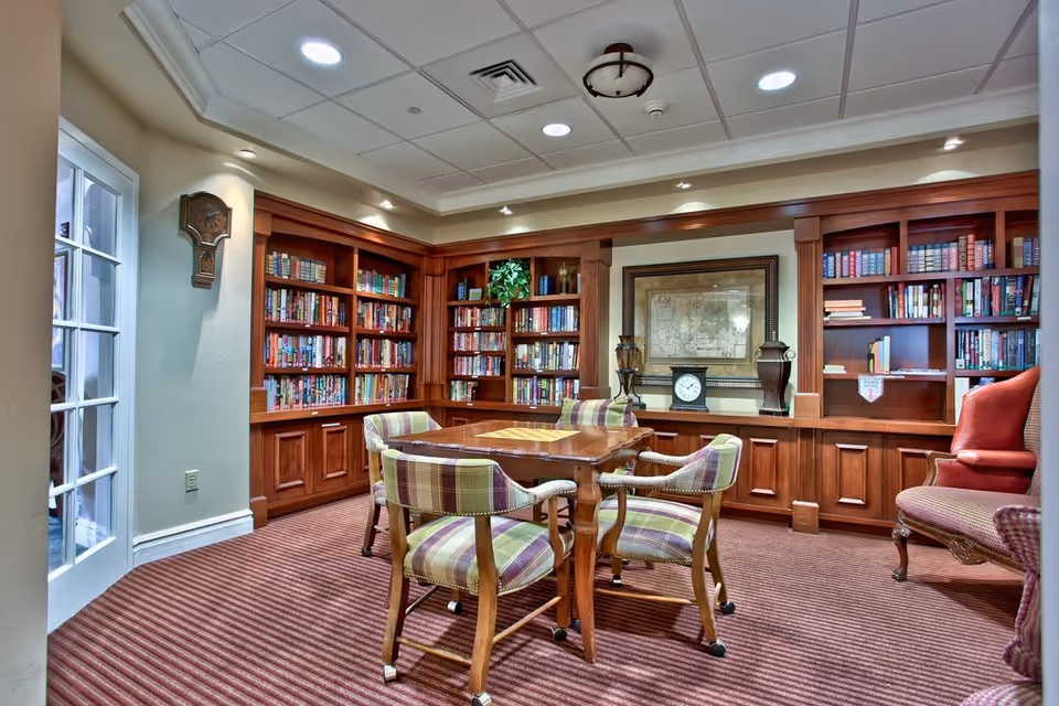 A cozy library room with wooden bookshelves filled with books, a wooden table with a built-in chessboard, and four plaid upholstered chairs around the table. The room has a carpeted floor, a framed map on the wall, decorative vases, and a clock on the shelf. There is also a red armchair on the right side and a wall clock on the left wall near a glass door.