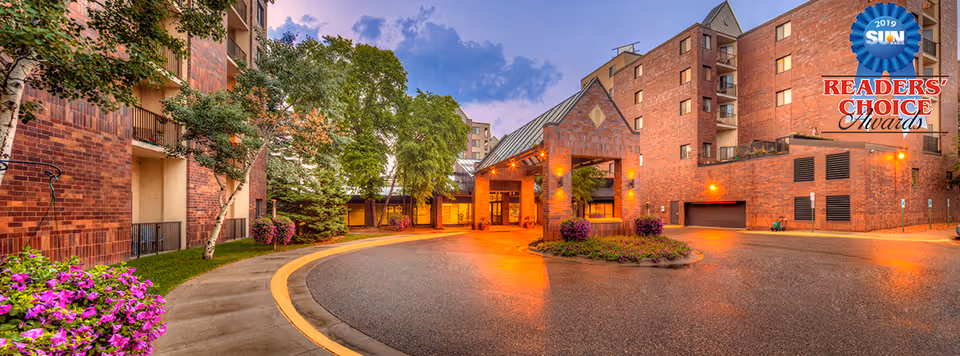 Entrance driveway of a multi-story brick building with landscaped greenery and flowers, illuminated by warm exterior lights during dusk. The building has balconies and a covered entrance area. A blue and gold badge in the top right corner reads '2019 SUN Readers' Choice Awards'.