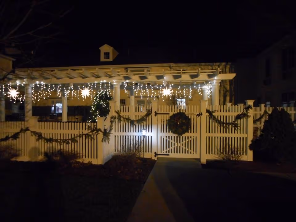 Night view of a white picket fence and gate decorated with garlands and a wreath, illuminated by hanging string lights and star-shaped lights, with a pergola and building in the background.