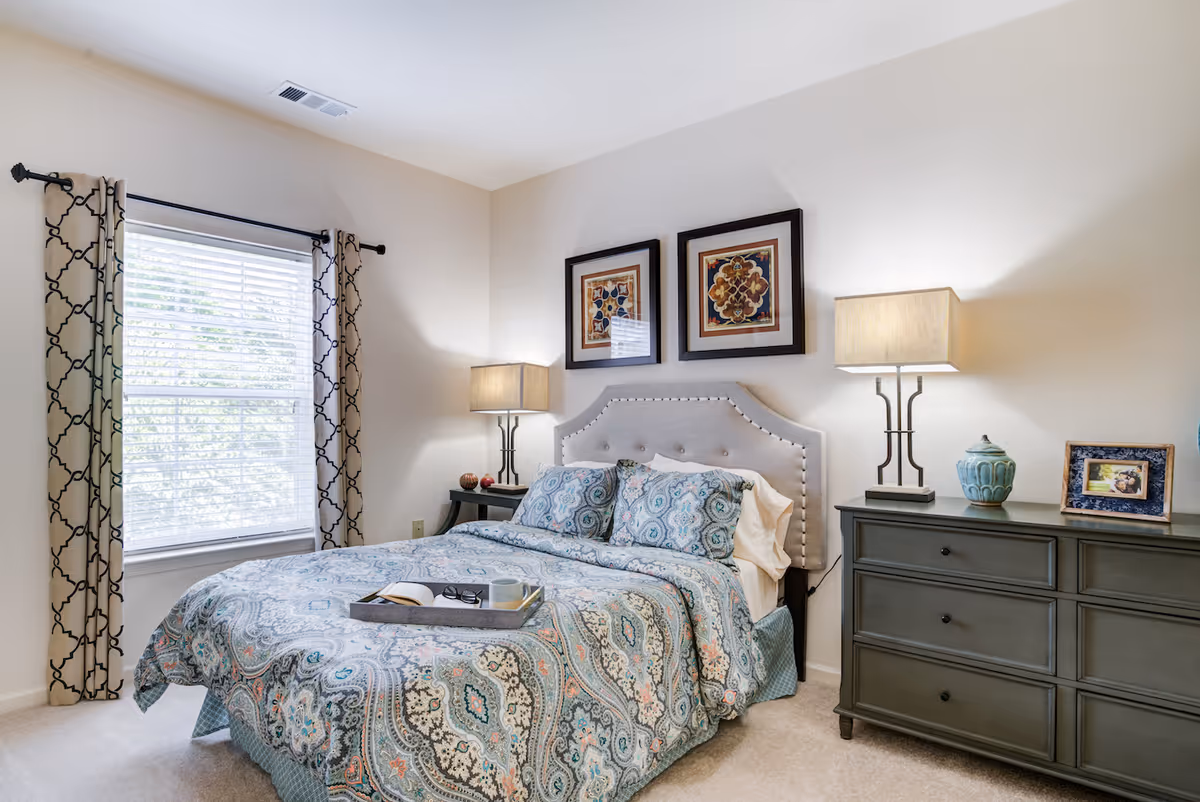 A neatly made bed with a patterned blue and beige comforter and matching pillows in a bedroom. The bed has a tufted headboard with nailhead trim. On either side of the bed are dark wood nightstands with lamps. Above the bed are two framed decorative artworks. A window with patterned curtains is on the left side of the room, allowing natural light to enter.