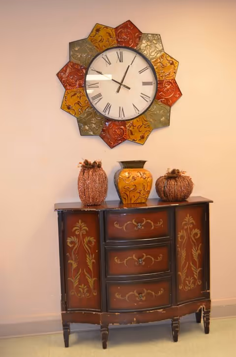 A decorative wall clock with Roman numerals and a colorful, flower-shaped frame hangs above a vintage wooden cabinet with ornate floral designs. On top of the cabinet are three decorative items: two pumpkin-shaped ornaments and a yellow vase with floral patterns.