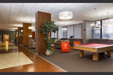 Interior view of a senior living facility common area featuring a pool table, orange armchairs, a sofa, potted plants, and modern hanging light fixtures. The space has a mix of carpet and wood flooring, with large windows allowing natural light to enter.