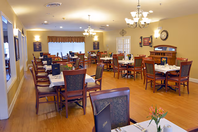 A well-lit dining room with multiple wooden tables and chairs arranged neatly. Each table is set with white tablecloths, menus, and small flower arrangements. The room has wooden flooring, beige walls, and decorative wall art. There are chandeliers hanging from the ceiling providing warm lighting.