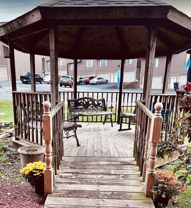 A wooden gazebo with a peaked roof and railings, containing a metal bench and two chairs with cushions. The gazebo is accessed by wooden steps and is surrounded by potted plants and flowers. In the background, there are parked cars and residential buildings.