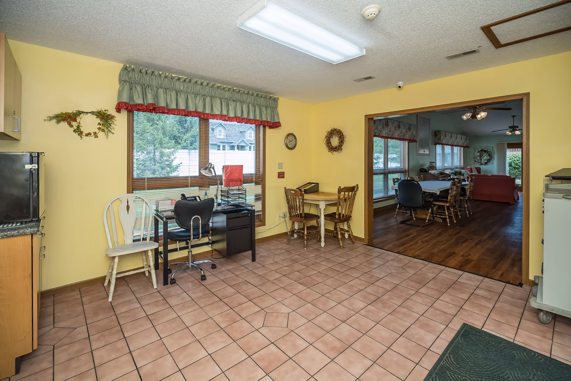 Interior view of a room with yellow walls and tiled floor featuring a small desk with a black office chair and a white wooden chair near a window with green and red valance curtains. There is a small round table with two wooden chairs against the wall. Through a large open doorway, a living room area with wooden flooring, multiple chairs, tables, and sofas is visible. The room is well-lit with ceiling lights and has decorative wreaths on the walls.