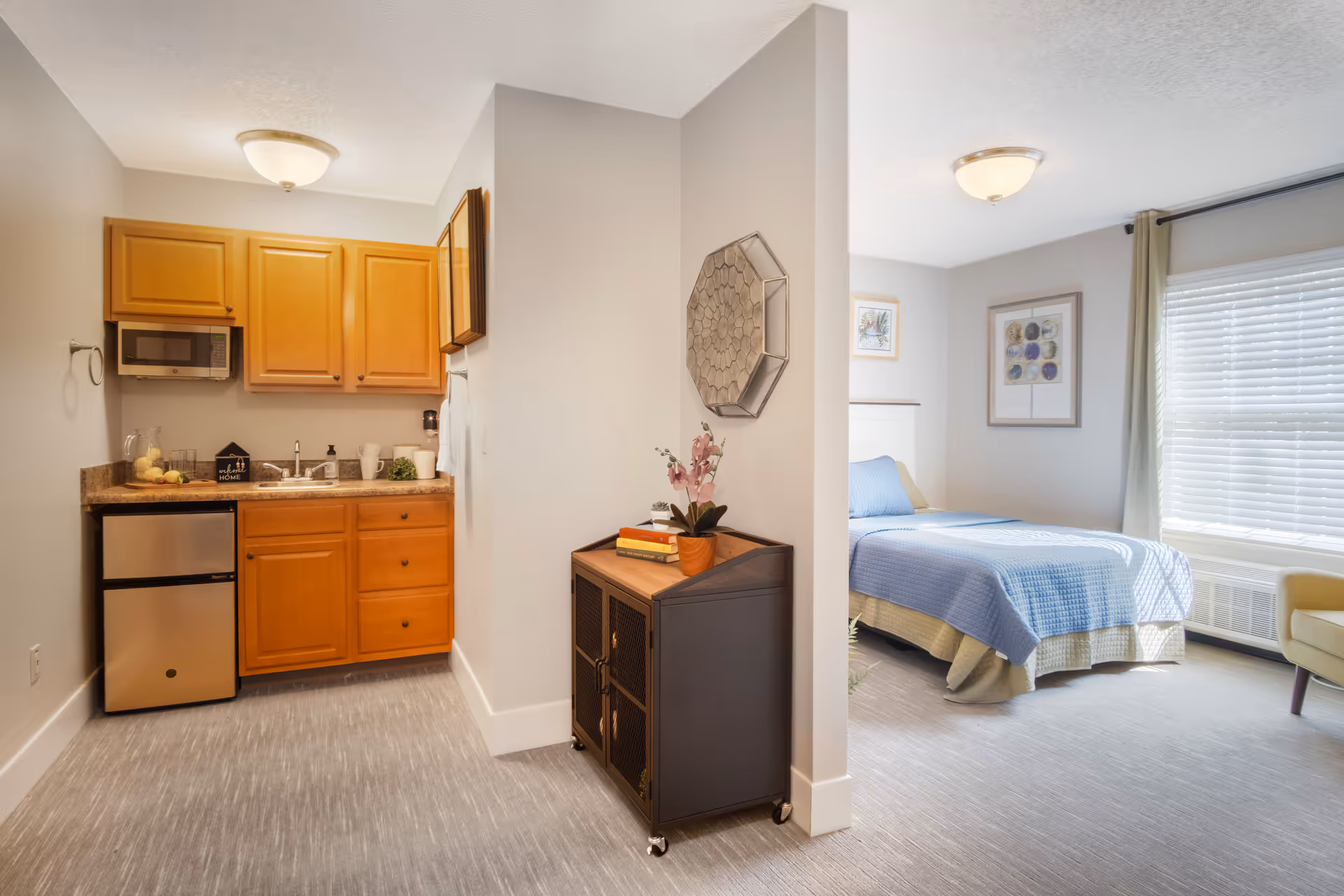 A bright and clean studio apartment featuring a small kitchenette with wooden cabinets, a mini refrigerator, microwave, and sink on the left. To the right, there is a cozy bedroom area with a bed covered in a blue quilt, a window with blinds and curtains, framed artwork on the walls, and a yellow armchair. A small cabinet with books and a potted plant is placed against a partial wall dividing the kitchenette and bedroom areas.
