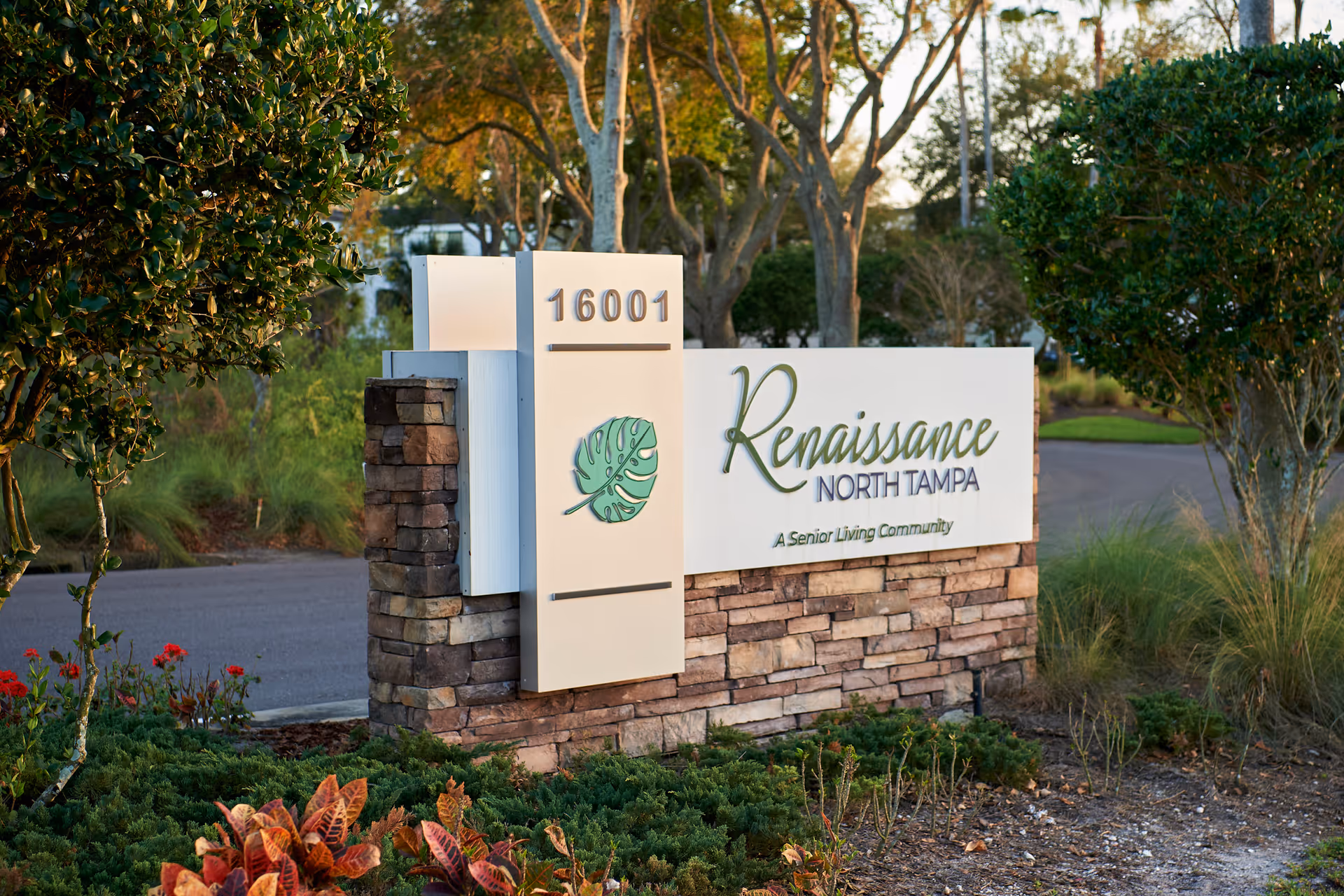 Outdoor view of a stone and white sign for Renaissance North Tampa, a senior living community, with the address number 16001. The sign is surrounded by greenery and trees with a road in the background.