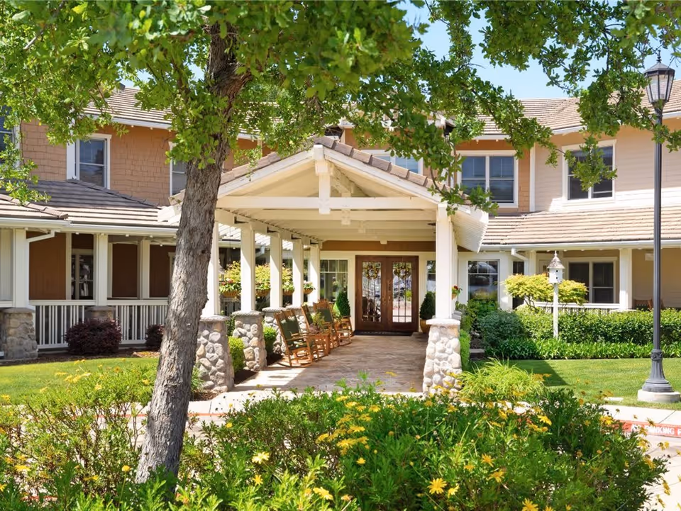 Entrance of a senior living facility with a covered porch supported by white pillars and stone bases. Several wooden rocking chairs are lined up under the porch. The building has beige siding and multiple windows. There is a tree and green bushes with yellow flowers in the foreground, and a lamppost on the right side.