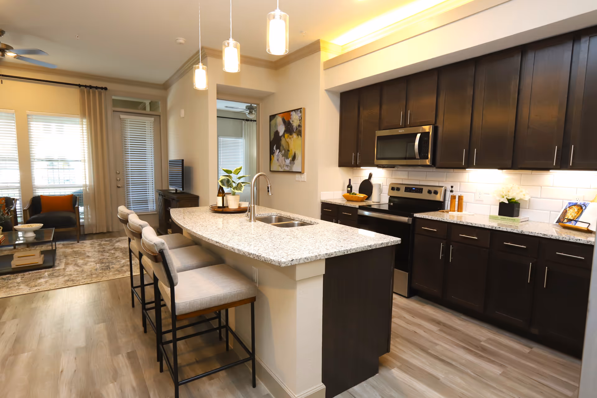 Modern kitchen with dark wood cabinets, stainless steel appliances including a microwave and stove, and a granite countertop island with a sink and three cushioned bar stools. The kitchen opens into a living area with a TV, armchairs, and large windows with curtains.