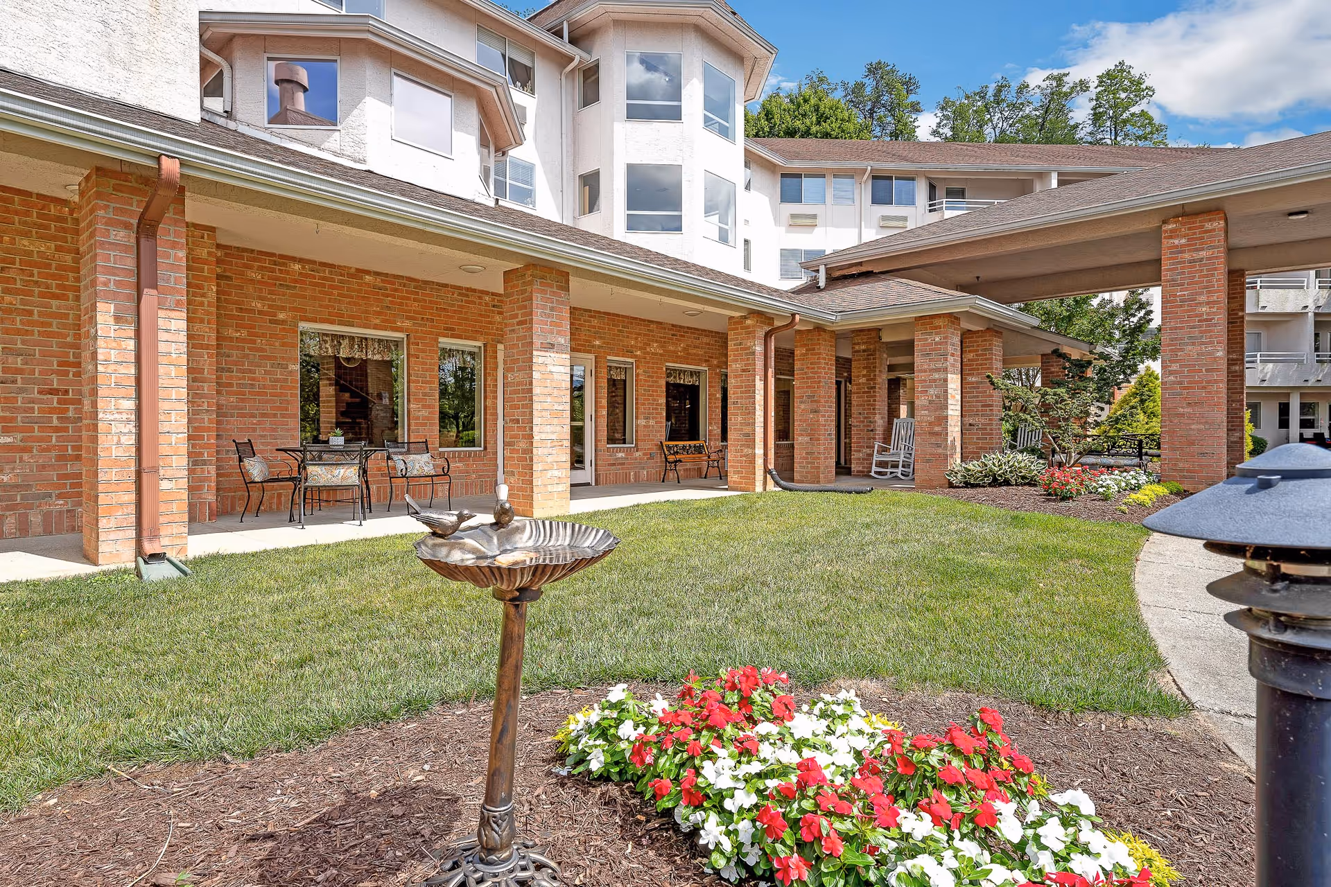 Brick senior living facility courtyard with a birdbath, colorful flowers, patio seating, and a covered entrance.